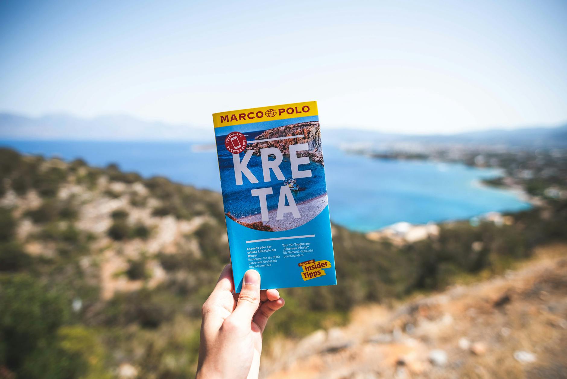 A person holds a travel guidebook overlooking a scenic coastline under a bright blue sky.