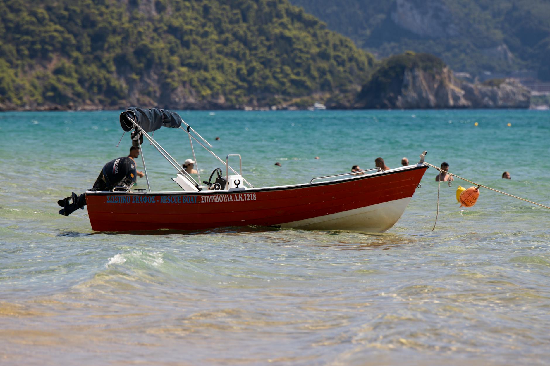 Serene scene of a rescue boat on a clear, turquoise beach in Greece with swimmers in the distance.