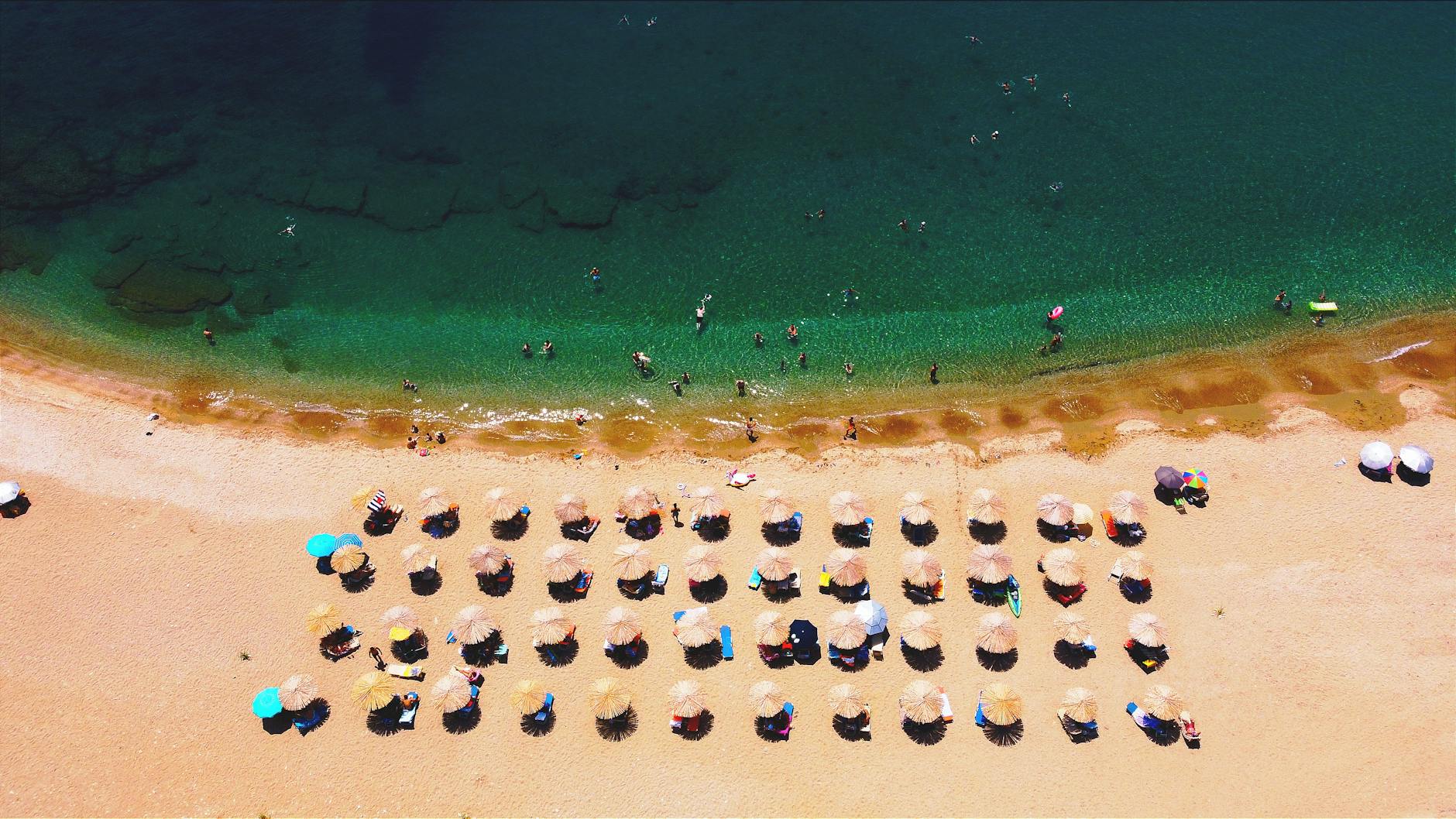 Aerial view of a sunny beach in Greece with umbrellas and swimmers enjoying the sea.